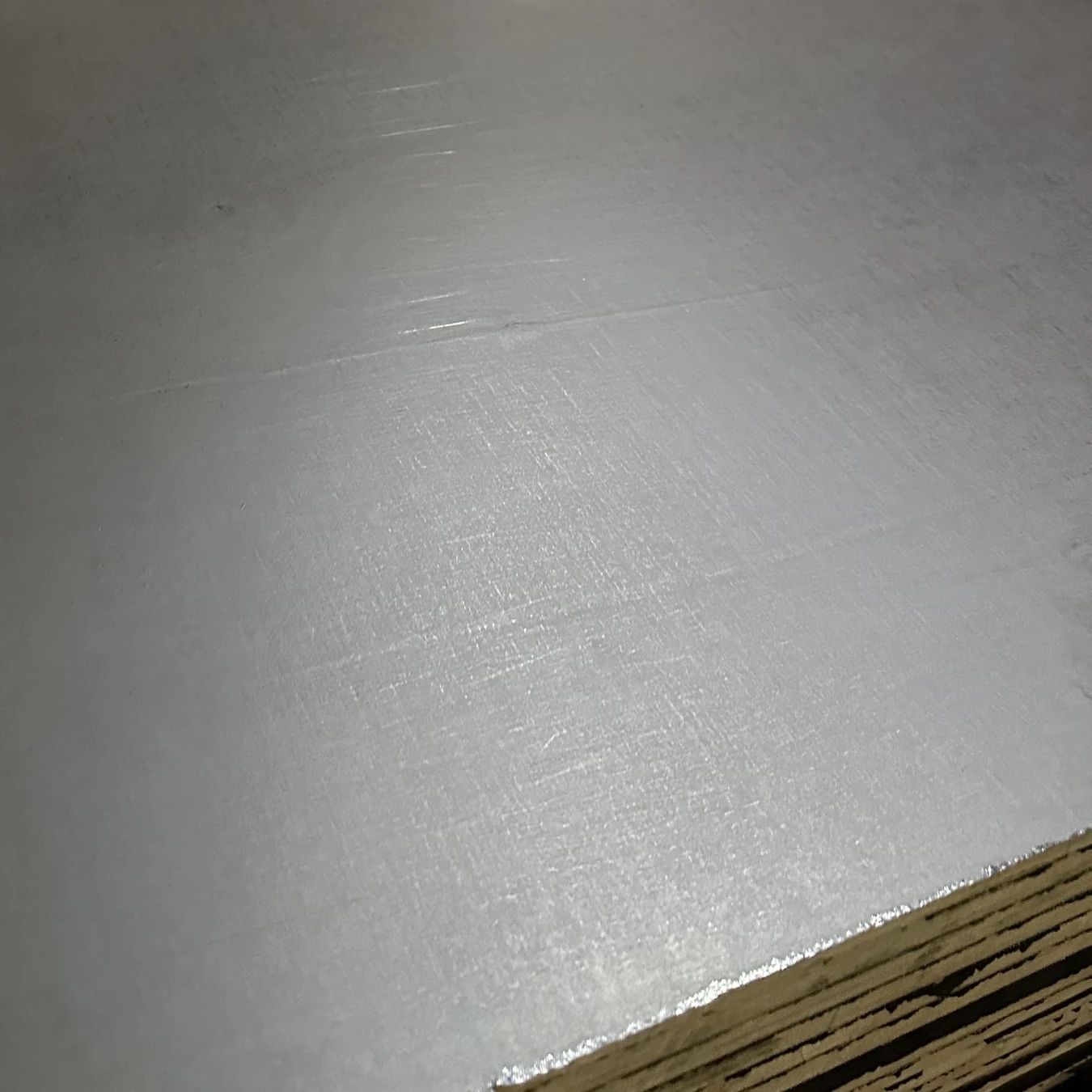 Film face of a plywood panel catching the light, fine woven texture, with its front edge and stacked edges below
